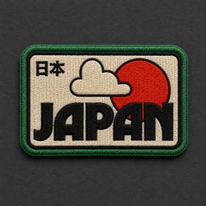 May include: A rectangular embroidered patch with a cream-colored background and a thick green border. It features the black text JAPAN, a white cloud, a red circle, and the black Japanese characters 日本, all rendered in a bold, stitched design.