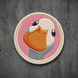 May include: A circular embroidered patch featuring a stylized goose head with a white face, a large orange beak, and dark blue eyes. The goose is set against a solid pink background, and the entire design is framed by a cream-colored stitched border.