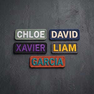 May include: Five rectangular embroidered name patches featuring the names CHLOE in white on sage green, DAVID in cream on navy blue, XAVIER in purple on black, LIAM in yellow on dark gray, and GARCIA in teal on burnt orange with stitched borders.