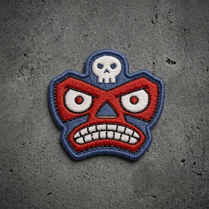 May include: An embroidered patch featuring a stylized mask design with a blue border, red face, and white details. The design includes a small white skull at the top, two large white eyes with red pupils, and a wide mouth showing white teeth.