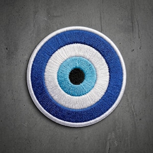 May include: A circular embroidered patch featuring a concentric eye design. The pattern consists of a solid black center, surrounded by rings of light blue, white, and a dark blue outer border, all rendered in dense, textured thread stitching.