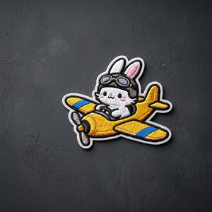May include: An embroidered patch featuring a white rabbit wearing brown aviator goggles and a matching cap, piloting a yellow airplane with blue stripes on the wings and a black propeller, all outlined in a thick white border.