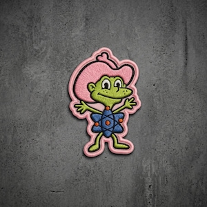 May include: An embroidered patch featuring a green cartoon character wearing a pink cowboy hat. The character has a blue atom-shaped emblem on its chest with orange dots, and is depicted with arms outstretched against a light pink border.