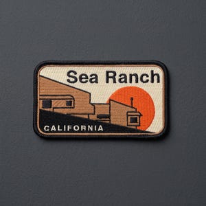 May include: A rectangular embroidered patch featuring a tan building silhouette against a cream background with a large orange sun. The text Sea Ranch is embroidered in black at the top, and the word CALIFORNIA is in white at the bottom.