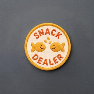 May include: A circular embroidered patch with a cream-colored background and a thick yellow border. The text SNACK DEALER is stitched in orange above and below two smiling, golden-yellow fish icons with three small orange dots between them.
