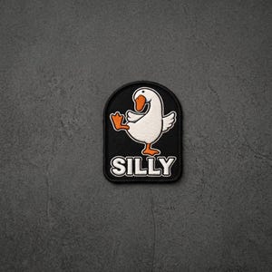 May include: A black embroidered patch with a rounded top edge featuring a white goose with an orange beak and orange feet standing on one leg. Below the goose, the word SILLY is embroidered in bold white capital letters with a black outline.