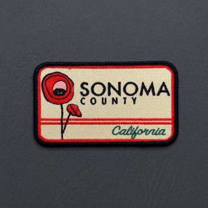 May include: A rectangular embroidered patch with a cream background and a black border. It features a red poppy flower, the black text SONOMA COUNTY, and the green text California. Two thin red horizontal lines run across the center of the patch.