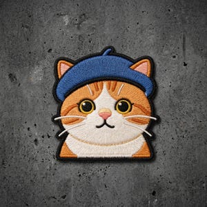 May include: An embroidered patch featuring a ginger and white cat wearing a blue beret. The cat has large yellow eyes, a pink nose, and white whiskers. The design is outlined in black thread, showcasing detailed stitching on the fur and hat.