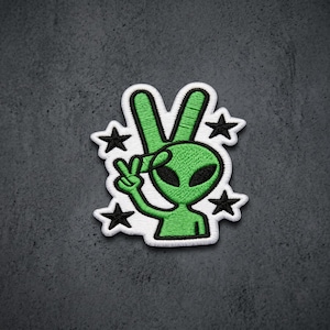 May include: An embroidered patch featuring a bright green alien character making a peace sign gesture. The alien has large black oval eyes and is surrounded by four small black embroidered stars. The entire design is outlined in white thread.