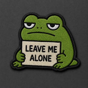 May include: An embroidered patch featuring a green frog with a grumpy expression holding a beige rectangular sign. The sign has black text that reads LEAVE ME ALONE. The entire design is outlined in thick black thread with detailed stitching.