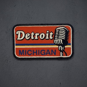 May include: A rectangular embroidered patch with an orange background and a dark blue border. It features the white text Detroit above the blue text MICHIGAN. A black and white vintage-style microphone graphic is positioned on the right side.