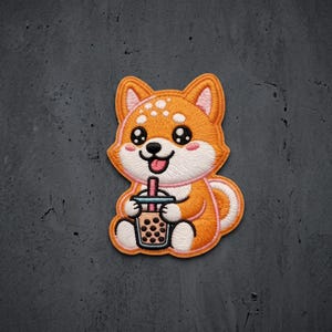 May include: An embroidered patch featuring a cartoon Shiba Inu dog with orange and white fur, holding a cup of bubble tea with a pink straw and dark brown tapioca pearls. The dog has large black eyes, a small black nose, and a pink tongue showing.
