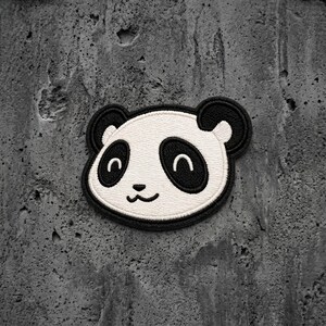 May include: An embroidered patch featuring the face of a panda. The design consists of a white face with black ears, black oval-shaped eye patches, a small black nose, and a curved black mouth, all outlined with a thick black stitched border.