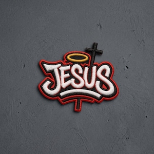 May include: An embroidered patch featuring the word JESUS in white lettering with a thick black outline. The design includes a yellow halo above the letters and a black cross integrated into the text, all bordered by a vibrant red stitched edge.