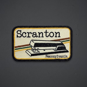 May include: A rectangular embroidered patch with a cream-colored background and a black border. It features the black text Scranton at the top and Pennsylvania at the bottom, with a black stapler graphic and diagonal brown, green, and yellow lines.