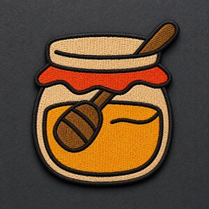 May include: An embroidered patch shaped like a glass jar filled with golden honey. The jar features a red fabric lid cover, a thick black border, and a brown wooden honey dipper resting inside the jar, angled toward the upper right corner.