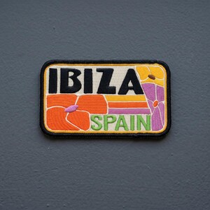 May include: A rectangular embroidered patch featuring the text IBIZA in bold black letters and SPAIN in green letters. The design includes orange, yellow, and purple floral motifs on a cream background with a thick black border edge.