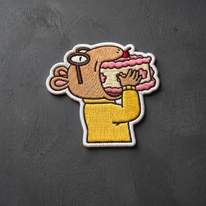 May include: An embroidered patch featuring a tan cartoon character wearing round glasses and a yellow sweater, holding a large slice of pink and white layer cake topped with a single red cherry. The design has a white border and stitching.