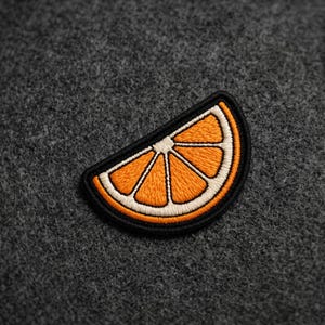 May include: An embroidered patch shaped like a slice of orange. The design features bright orange segments separated by white lines, all enclosed within a thick black border. The stitching is dense and textured, creating a clear fruit motif.