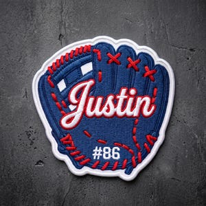May include: A blue embroidered patch shaped like a baseball glove with red stitching details. The patch features the white script name Justin across the center and the white text #86 near the bottom, all outlined in a thick white border.