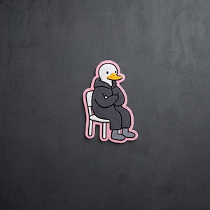 May include: A patch featuring a white duck with a yellow beak wearing a dark gray hooded sweatshirt and dark gray pants, sitting on a white chair. The entire figure is outlined in a thick, light pink border against a dark gray background.
