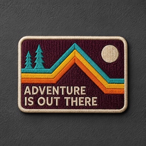 May include: A rectangular embroidered patch with a dark purple background featuring a mountain range design in teal, yellow, and orange stripes. Two teal pine trees stand on the left, a cream circle represents the sun, and the text ADVENTURE IS OUT THERE is stitched in cream.