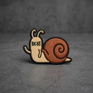 May include: An embroidered patch featuring a cartoon snail with a beige body and a brown spiral shell. The snail has a grumpy facial expression with green eyes and is outlined in thick black thread, giving it a distinct and bold appearance.