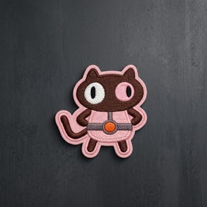 May include: An embroidered patch featuring a dark brown cat character with a white eye and a pink eye. The cat wears a light pink outfit with a dark gray belt and a central orange circular emblem. The entire design is outlined in light pink thread.