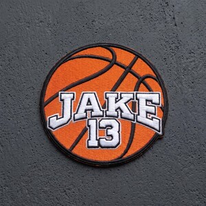 May include: A circular embroidered patch featuring an orange basketball design with black stitching details. The text JAKE 13 is embroidered in white with a black outline across the center of the basketball graphic in a bold, block-style font.