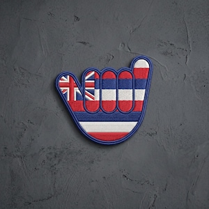 May include: An embroidered patch in the shape of a shaka hand gesture, featuring the red, white, and blue horizontal stripes of the Hawaii state flag, with the Union Jack pattern visible in the upper left corner of the thumb and index finger.