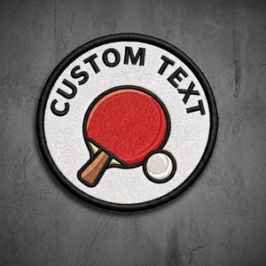 May include: A circular embroidered patch with a white background and a thick black border. The design features a red table tennis paddle with a brown handle and a white ball. The words CUSTOM TEXT are embroidered in black along the top curve.