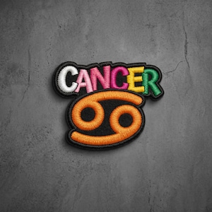 May include: An embroidered patch featuring the word CANCER in white, pink, magenta, yellow, and green thread. Below the text is the orange Cancer zodiac symbol, which consists of two interlocking circles, all outlined in thick black thread.