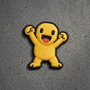 May include: An embroidered patch featuring a yellow cartoon character with a rounded head, black square eyes, and a wide open mouth with a red tongue. The character has raised arms and legs, outlined in black thread against a textured gray background.