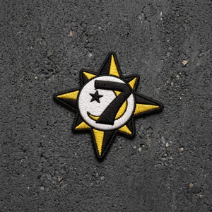May include: An embroidered patch featuring a central white circle with a black number 7 and a small black star. The circle is surrounded by a yellow and black starburst shape with eight points. The design uses bold black, white, and yellow thread.