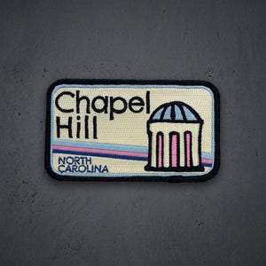 May include: A rectangular embroidered patch with a cream background and a black border. It features the text Chapel Hill and North Carolina in black, alongside a graphic of a domed building with light blue and pink accents and horizontal stripes.