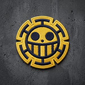 May include: A circular embroidered patch featuring a stylized yellow and dark blue skull emblem. The design includes two dark blue circular eyes, a small dot for a nose, and a wide grin with vertical bars, all set against a yellow gear-like border.