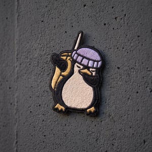 May include: An embroidered patch featuring a penguin wearing a light purple beanie and a black face mask. The penguin is holding a tan-colored beverage cup with a straw. The design uses black, white, tan, and purple thread on a dark background.