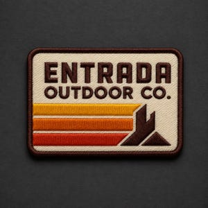 May include: A rectangular embroidered patch with a cream background and a dark brown border. The text ENTRADA OUTDOOR CO. is stitched in dark brown. Below the text are three horizontal stripes in yellow, orange, and red, ending in a brown mountain icon.
