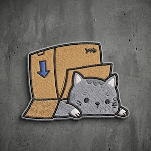 May include: An embroidered patch featuring a gray cat with white ears and paws peeking out from a tan cardboard box. The box has a blue downward-pointing arrow and a small black fish skeleton icon printed on the side, all outlined in black thread.