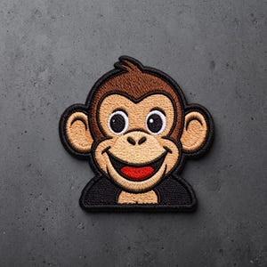 May include: An embroidered patch featuring a cartoon monkey face with brown fur, large ears, a tan muzzle, and a wide smile showing a bright red tongue. The design is outlined in thick black thread, creating a bold, defined shape for the character.