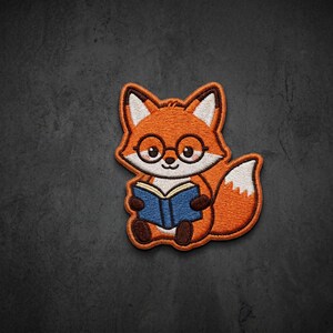 May include: An embroidered patch featuring a cartoon fox with orange fur, white accents, and brown paws. The fox wears round black glasses and is holding an open blue book. The design has a thick brown border and detailed stitching throughout.