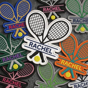 May include: A collection of embroidered patches featuring two crossed tennis rackets and a yellow tennis ball. Each patch displays the name RACHEL in bold capital letters across a horizontal bar. The patches come in various colors including blue, orange, green, pink, and white.