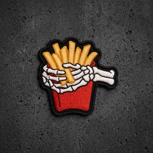 May include: An embroidered patch featuring a red carton filled with golden yellow french fries. A white skeleton hand is wrapped around the carton, gripping the fries. The design is outlined in black thread, creating a distinct graphic shape.