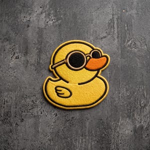 May include: A yellow embroidered patch in the shape of a duck wearing round black sunglasses. The duck has a bright orange beak and is outlined in a thick black border, featuring detailed stitching throughout the entire yellow body shape.