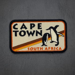 May include: A rectangular embroidered patch with a cream background, a black border, and the text CAPE TOWN in black. Below, the text SOUTH AFRICA appears in orange. Two black and white penguins stand over diagonal brown and orange stripes.