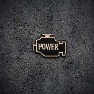May include: A black embroidered patch shaped like an engine block with a light beige border and the word POWER stitched in matching light beige thread across the center. The design features a stylized mechanical outline with clear lettering.
