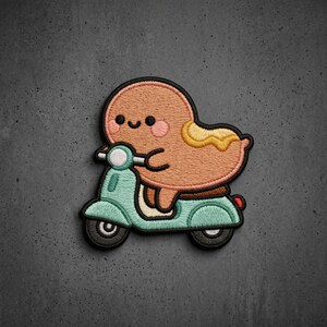 May include: An embroidered patch featuring a tan, rounded character with a smiling face and pink cheeks riding a light teal scooter. The character has a yellow, wavy shape on its back, and the entire design is outlined in black thread.