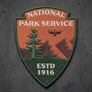 May include: A shield-shaped embroidered patch featuring an orange background with the white text NATIONAL PARK SERVICE. It depicts a green pine tree, brown mountains, and a bird silhouette. The bottom green section contains the white text ESTD 1916.