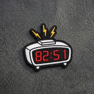 May include: An embroidered patch shaped like a digital alarm clock with a white border and black center. The display shows the red numbers 82:51. Three yellow lightning bolt icons extend from the top of the clock, indicating an alarm.