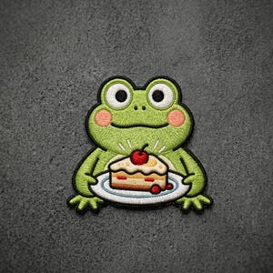 May include: An embroidered patch featuring a light green frog with large white eyes, black pupils, and pink cheeks. The frog is holding a white plate with a slice of cake topped with a red cherry. The patch has a thick black border outline.